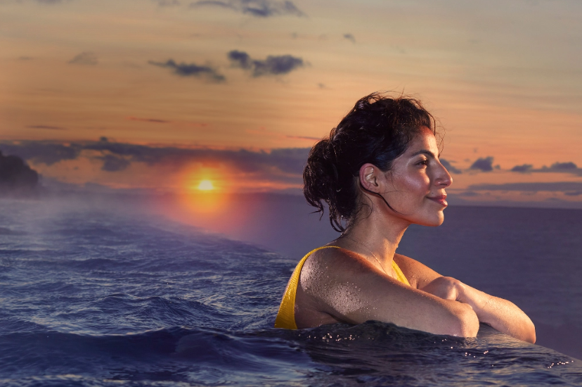 Woman in pool overlooking another body of water with a sunset and mountains in the background