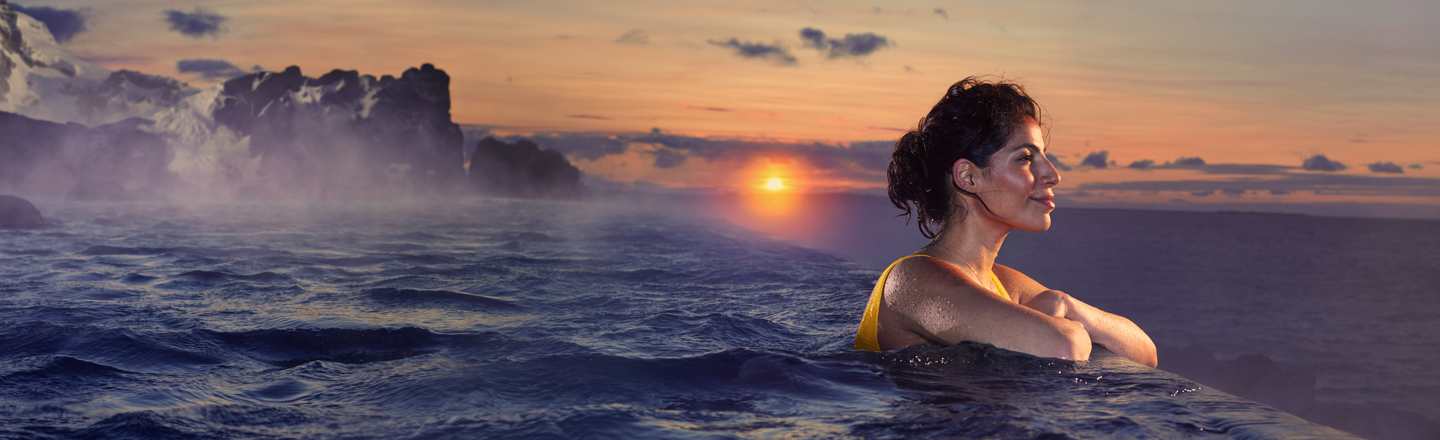 Woman in pool overlooking another body of water with a sunset and mountains in the background