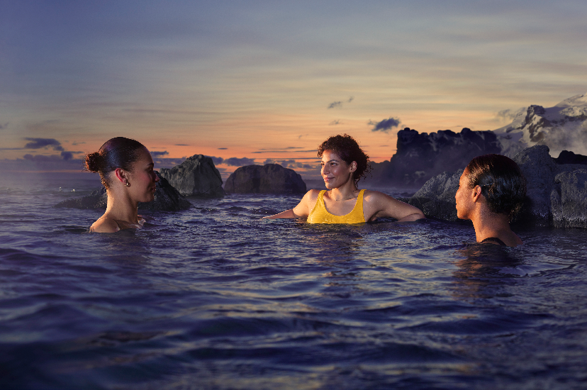 Three women smiling at each other in a body of water with rocks and mountains around them